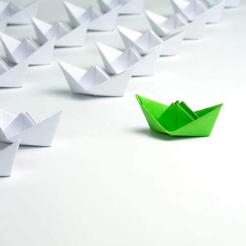 Leadership concept with Green paper ship standing out from the group of white ships on white background.